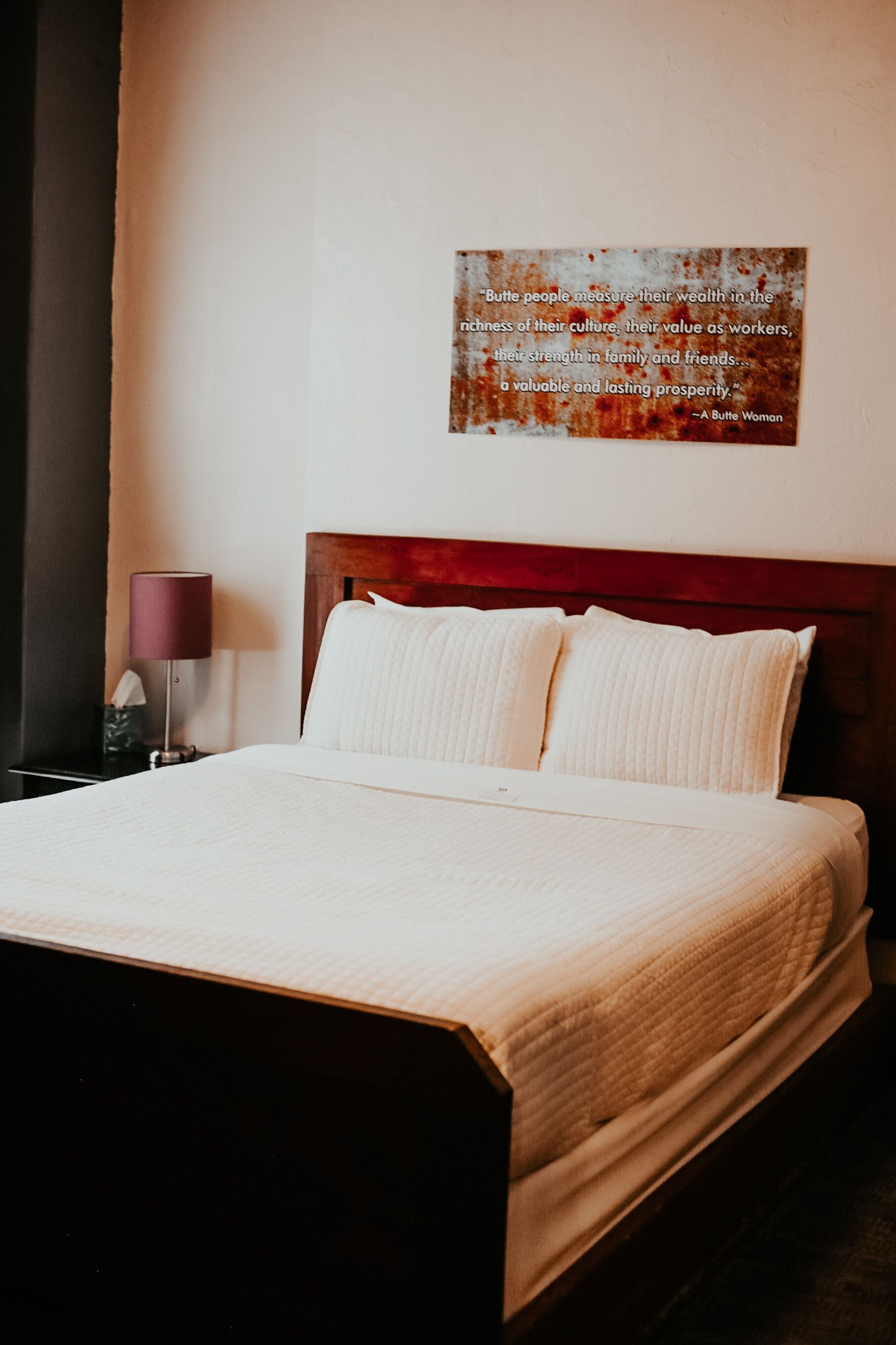 A neatly made bed with a wooden headboard, a bedside lamp, and a wall-mounted quote plaque.