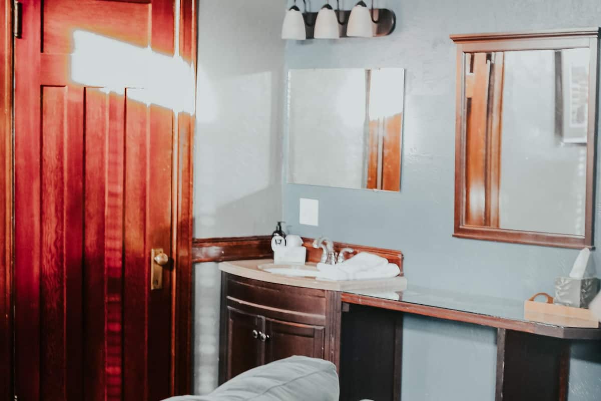 A room with a wooden door, a gray sofa, a wooden table, a small cabinet, a mirror, and a countertop with a sink and towel.