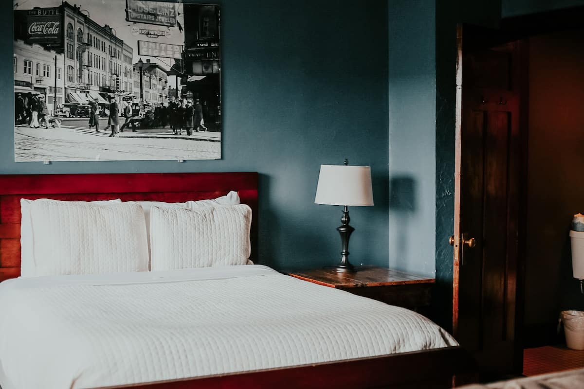 A hotel room with a large bed, a nightstand with a lamp, a wall-mounted picture of a street scene, and a wooden door.
