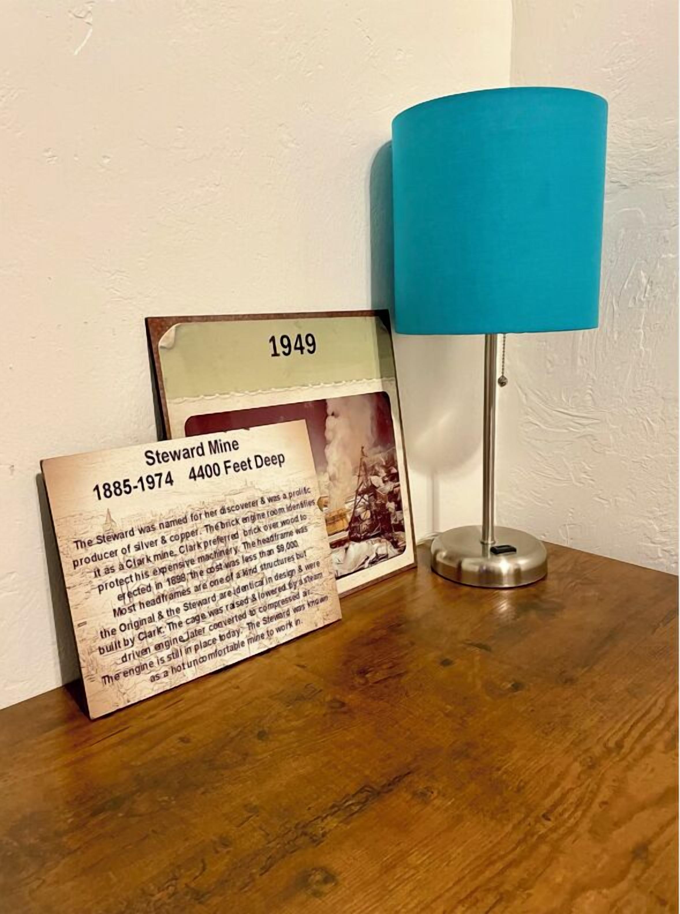 Un documento histórico enmarcado con el título 'Steward Mines 1885-1974 4400 Feet Deep' sobre una superficie de madera, junto a una lámpara azul.