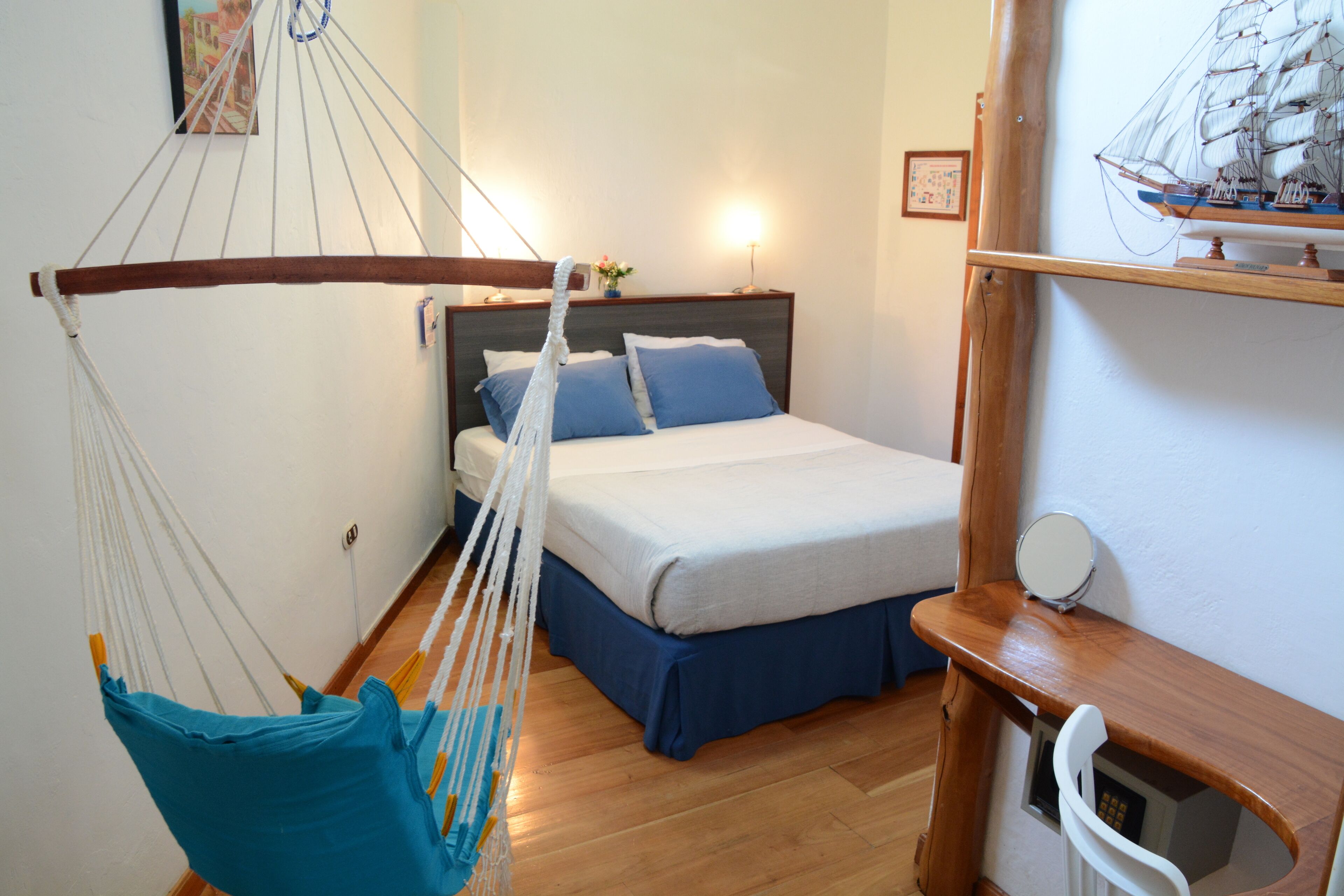 A bedroom with a bed, a hammock, a wooden shelf with a ship model, and a wall-mounted mirror.