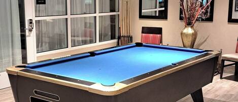 A pool table in a room with a large window, a vase with branches, and a framed mirror on the wall.