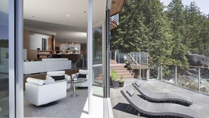 Una vivienda moderna con entrada de vidrio, una sala de estar con sofá y mesa de centro, y un balcón con sillones de descanso y vista a árboles.