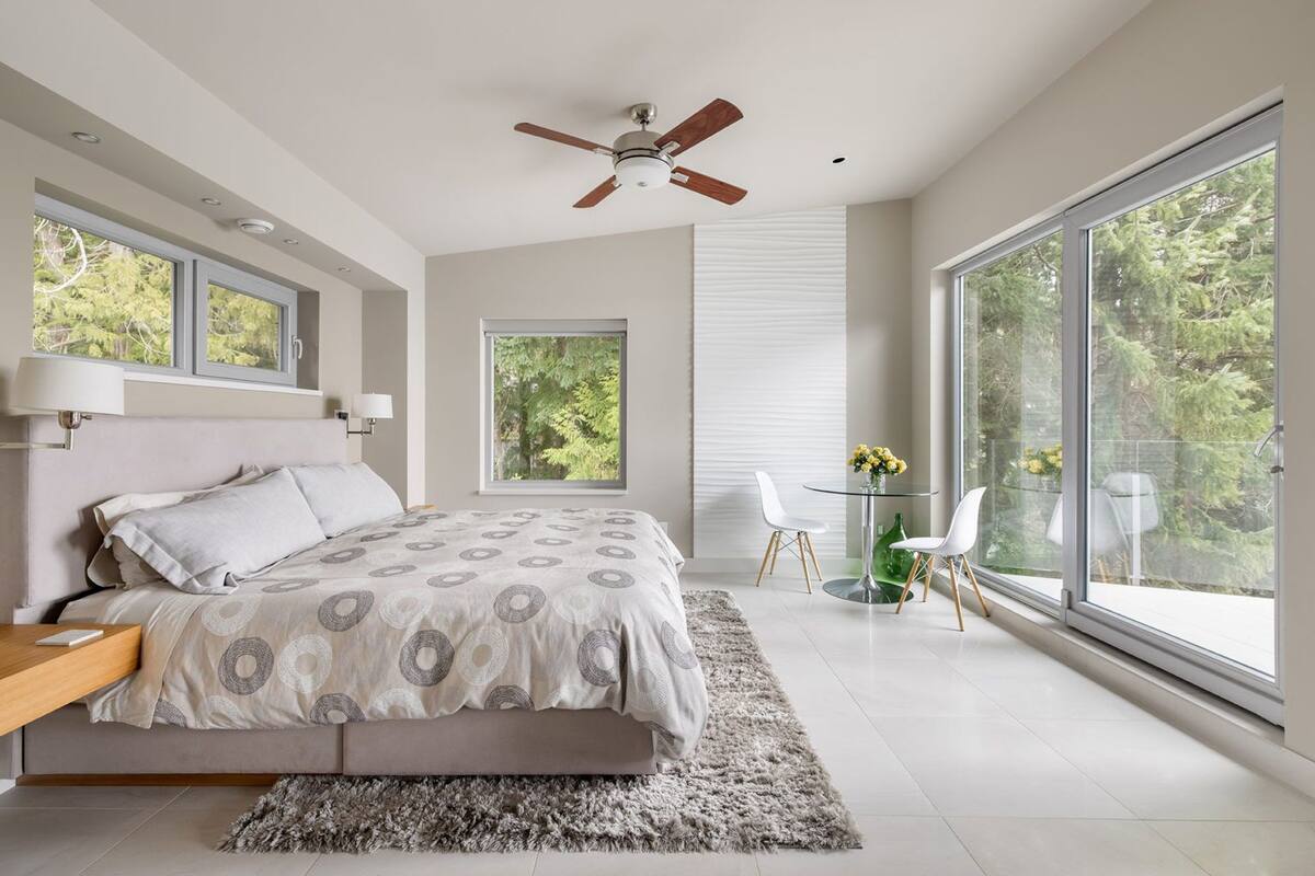 A modern bedroom with a large bed, a ceiling fan, and a view of trees through large windows.