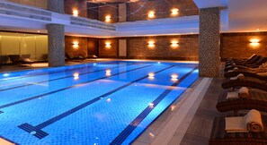 Une grande piscine intérieure avec des lignes de nage clairement délimitées, entourée de chaises longues et dotée d’un haut plafond avec des poutres apparentes.