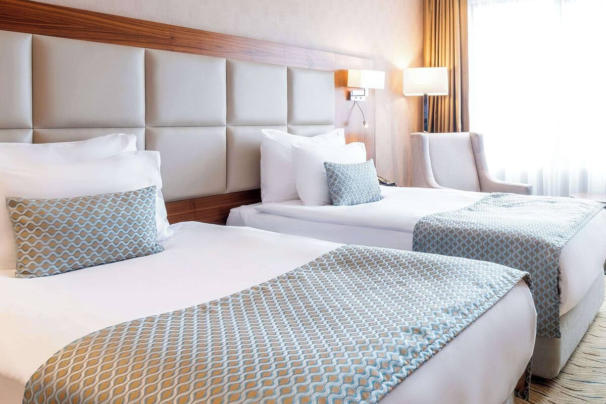 A hotel room with two beds, a wooden headboard, a lamp, and a view of the outdoors.