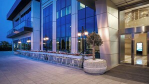 Un hôtel moderne avec une façade attrayante, une longue terrasse aménagée, de grandes fenêtres et une entrée bien éclairée.