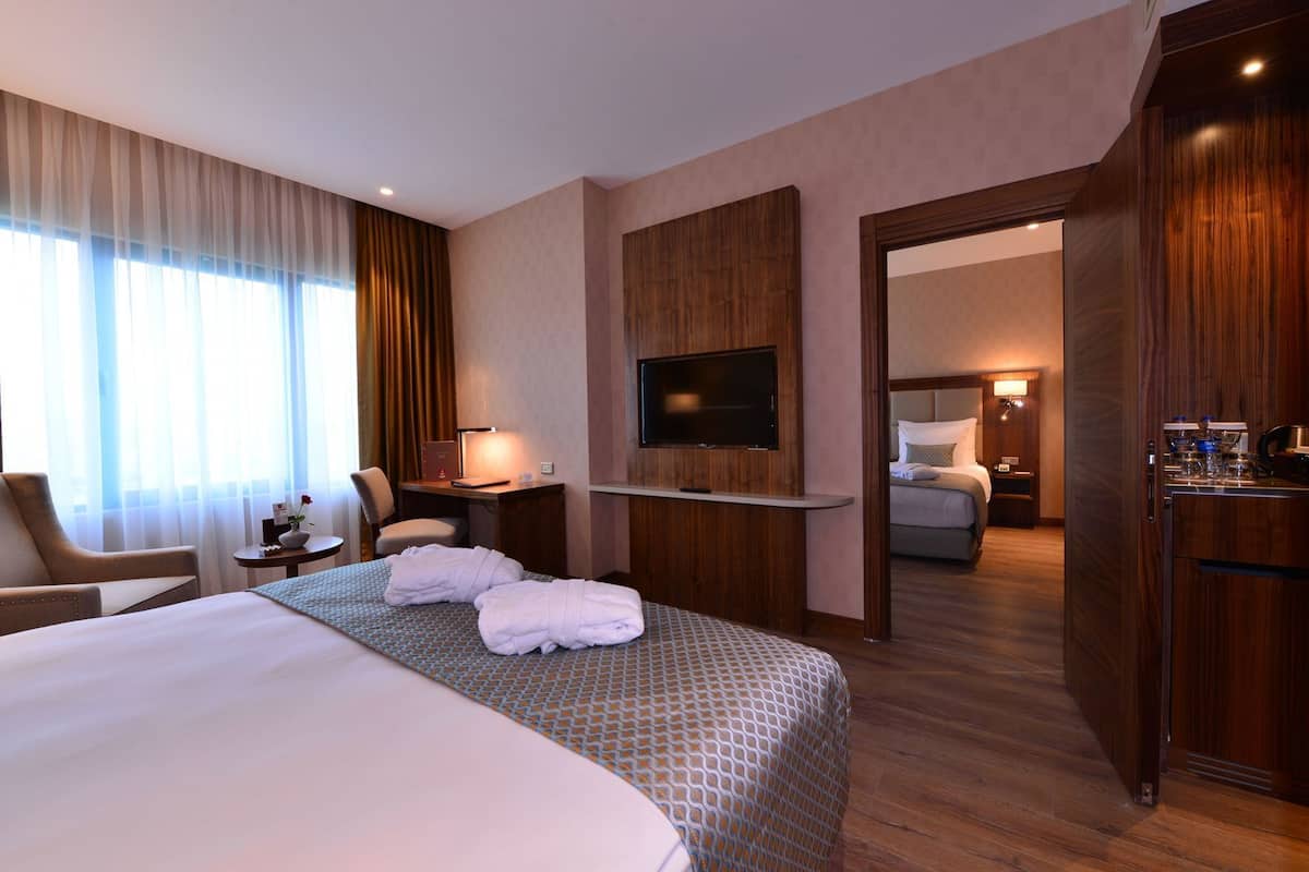 A hotel room with a large bed, a desk with a flat-screen TV, a chair, a small table with a lamp, and a minibar.