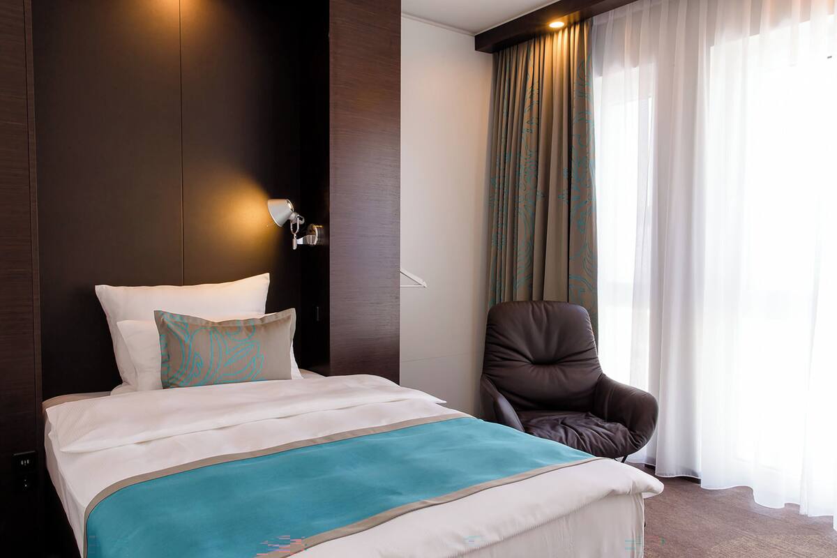 A hotel room with a bed, a chair, a wall-mounted lamp, and sheer curtains.