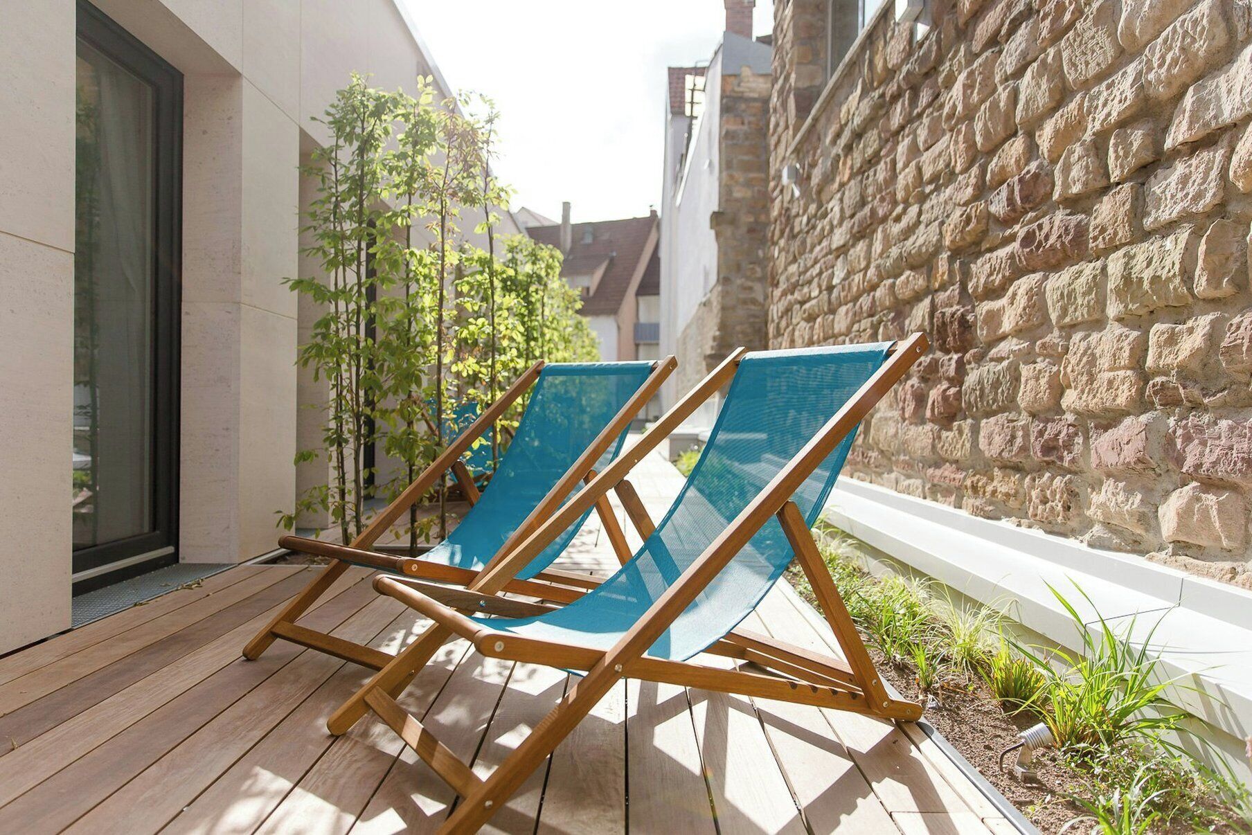 Zwei Holz-Loungesessel mit blauen Netzhocklehnen auf einer Holzterrasse neben einer Steinmauer.