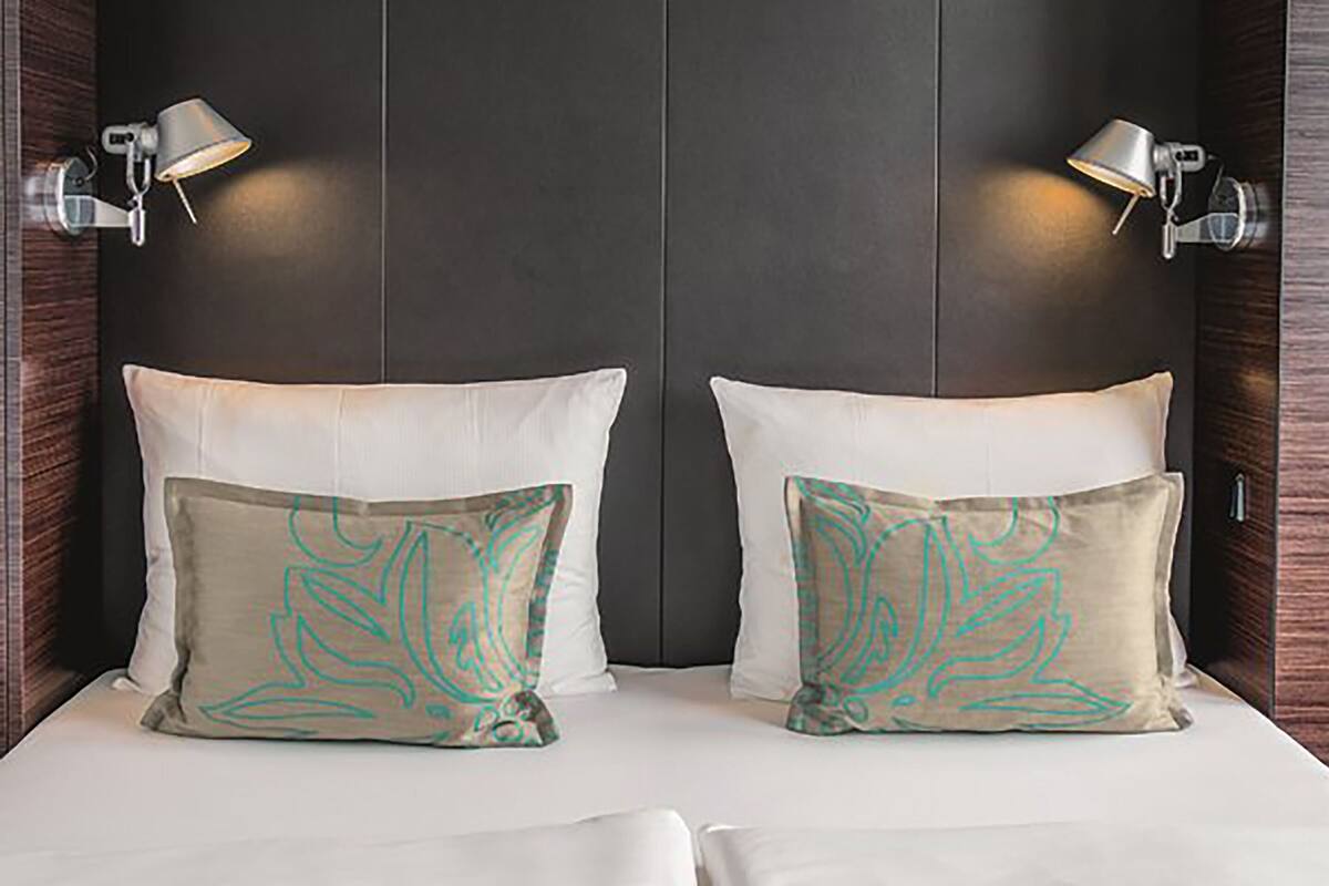 A double bed with two pillows, each with a unique design, and two wall-mounted lamps on either side.