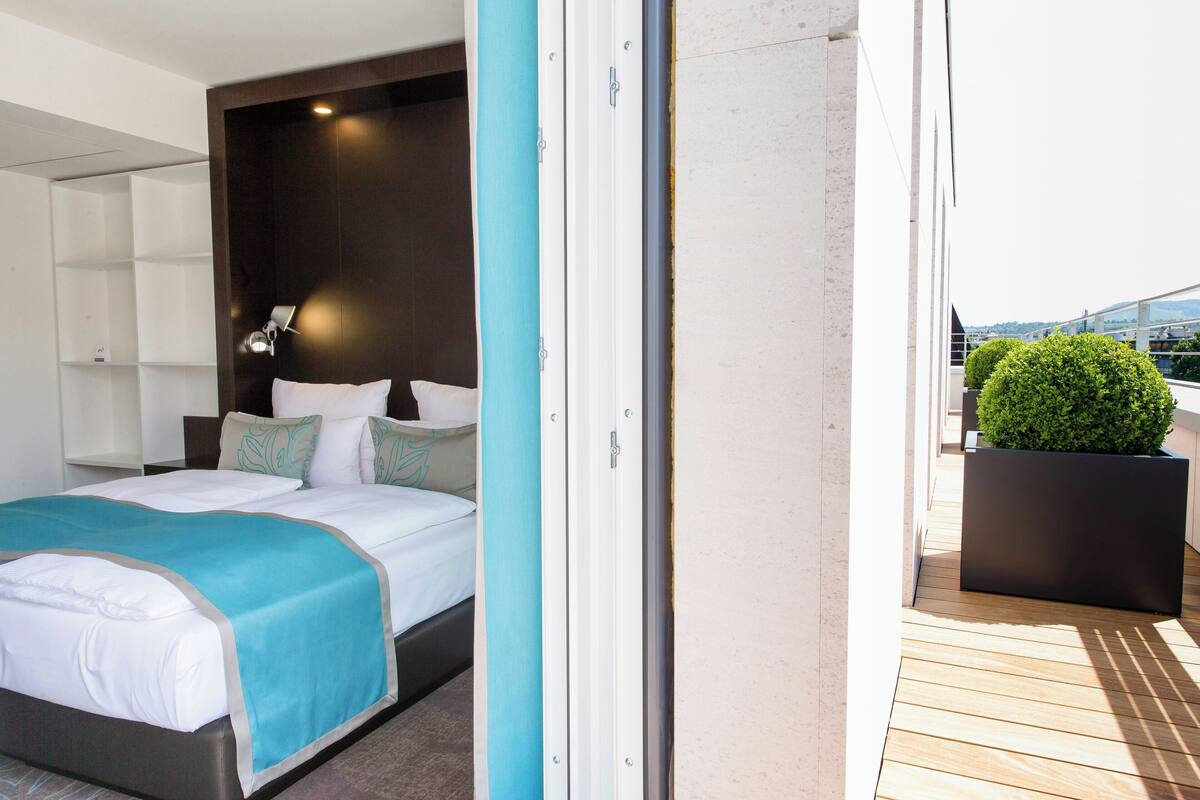 A modern hotel room with a bed, a nightstand, a wall-mounted lamp, and a view of the outdoors.
