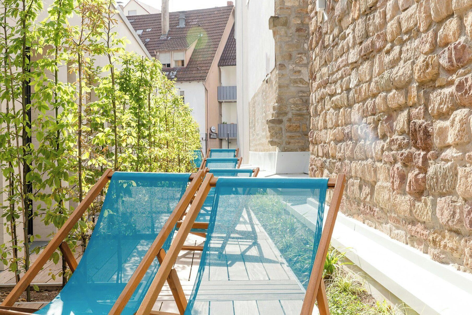 A row of blue lounge chairs with wooden frames, aligned along a pathway with a stone wall on one side and a row of trees on the other.