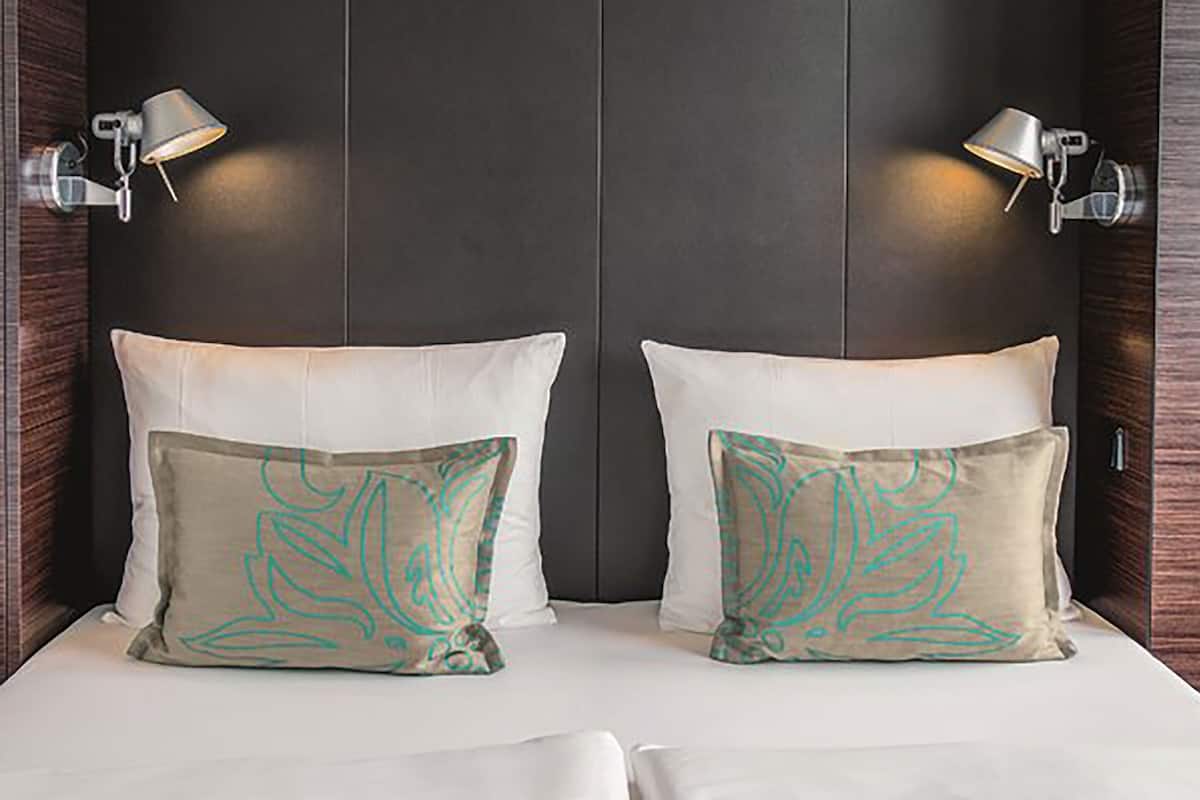 A double bed with two pillows, each with a unique design, and two wall-mounted lamps on either side.