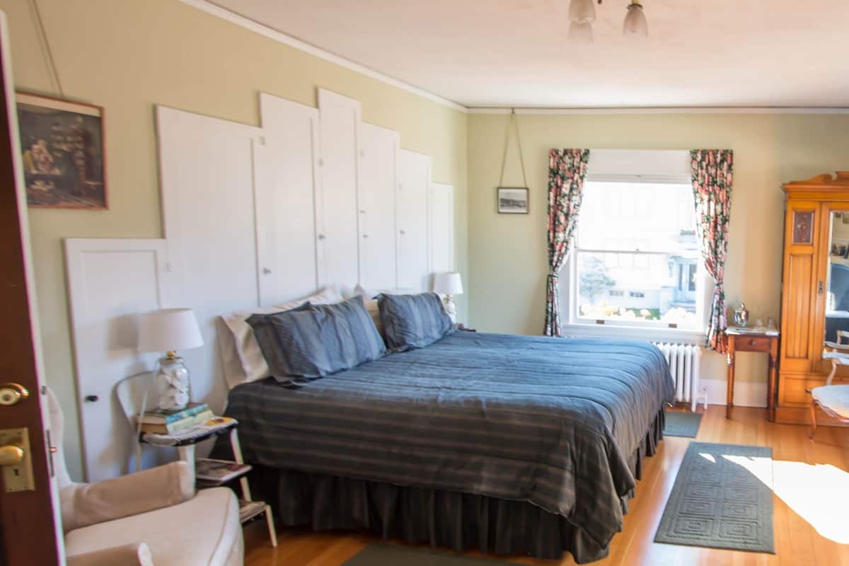 A bedroom with a large bed, a chair, a nightstand, a window with curtains, and a radiator.