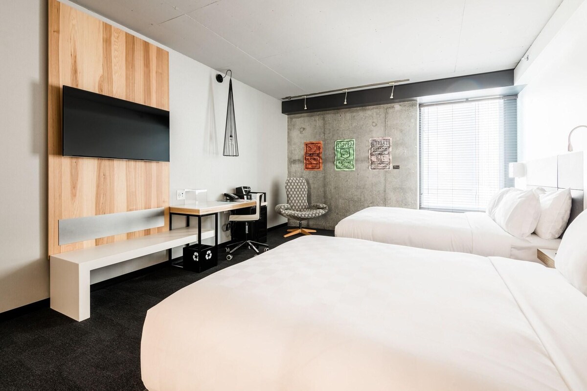 A modern hotel room with a large bed, a desk with a chair, a TV mounted on a wooden panel, and a window with blinds.