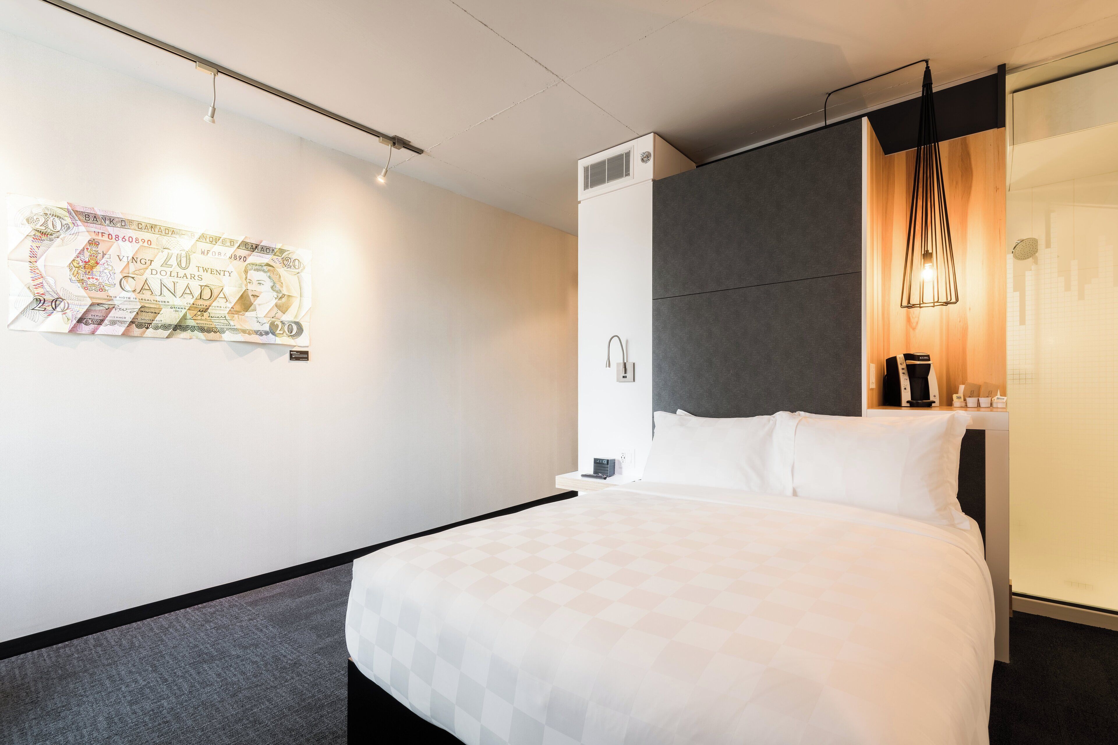 Una habitación de hotel con una cama, aire acondicionado de pared, una mesita de noche con lámpara y una obra de arte en la pared.