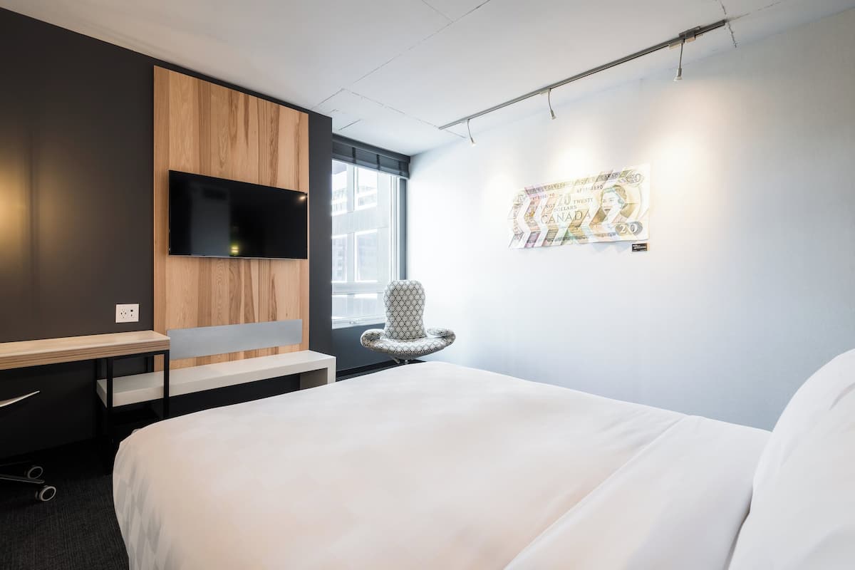 Ein modernes Hotelzimmer mit einem großen Bett, einem Flachbildfernseher an einer Holzverkleidung, einem Schreibtisch mit Stuhl sowie einem Wandbild.