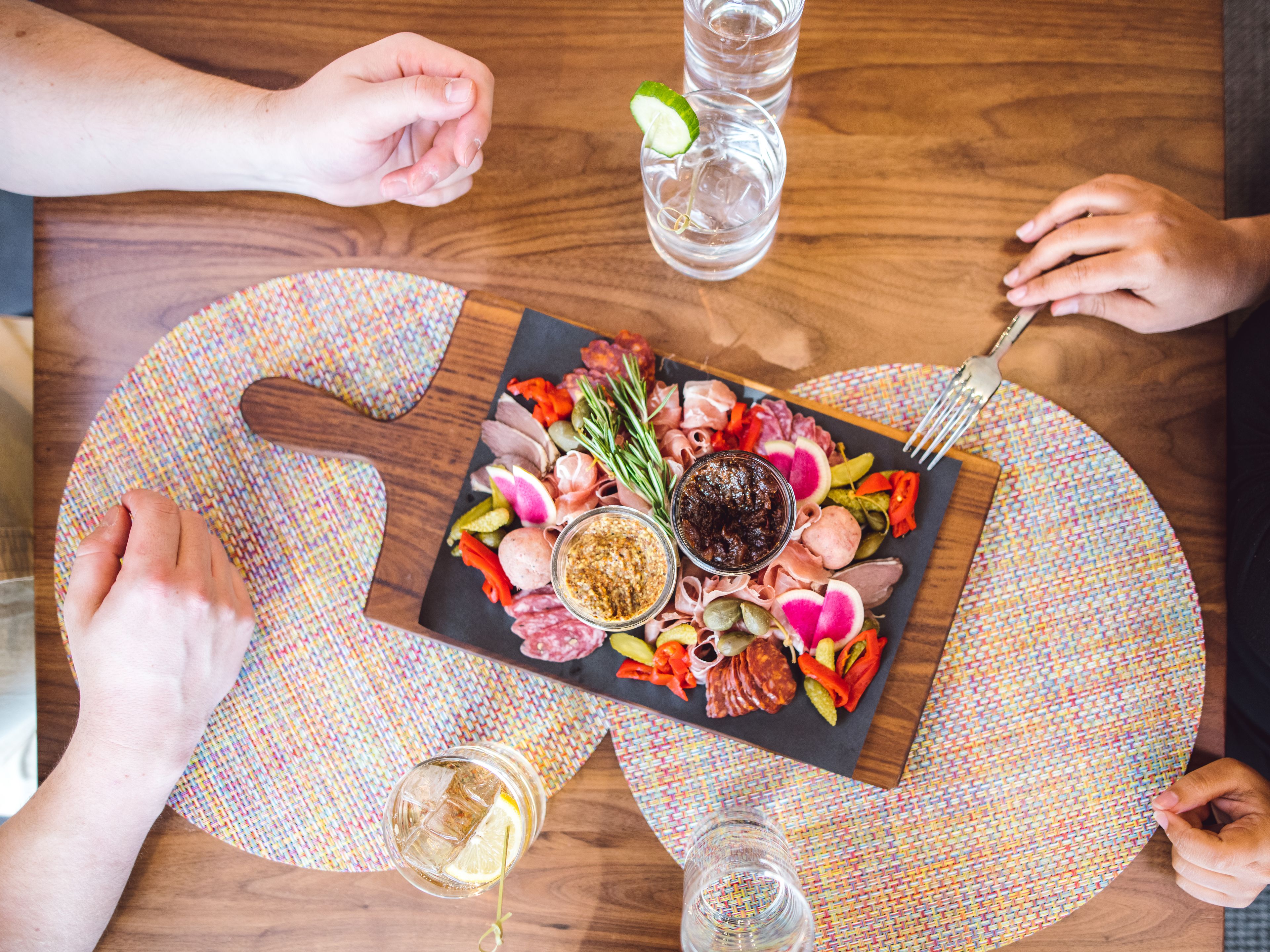 A round wooden table with a colorful woven placemat, a black slate board with various meats, vegetables, and condiments, and two glasses of water.