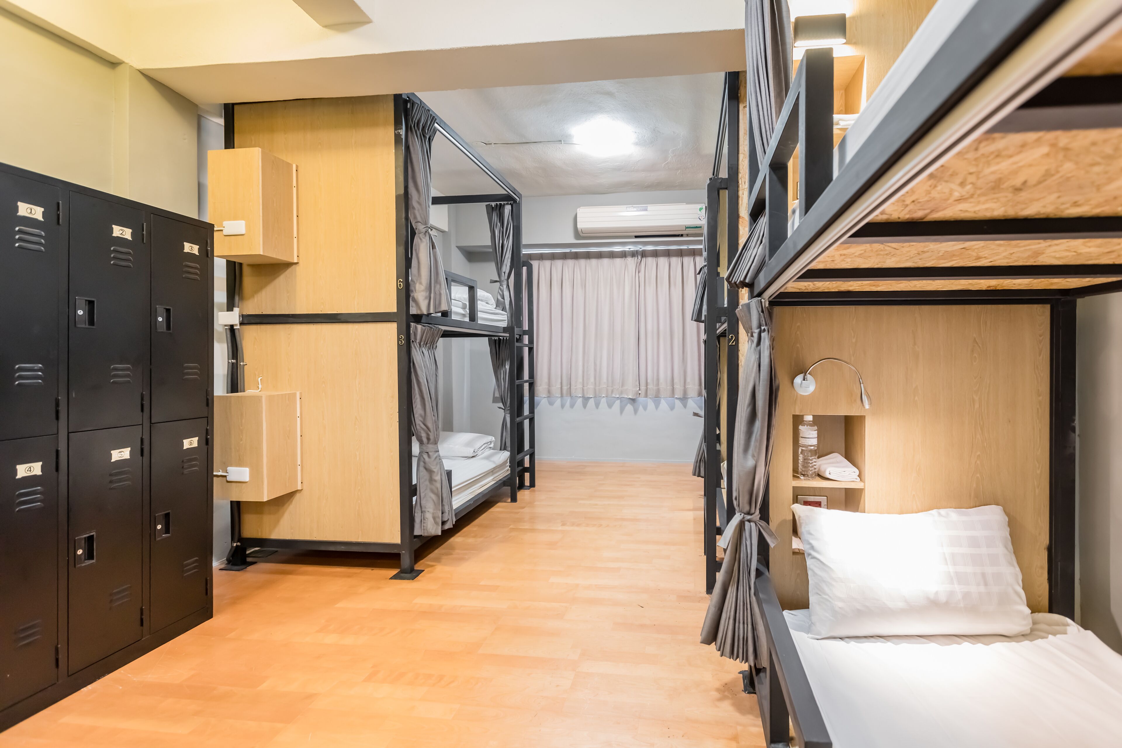A compact hotel room with bunk beds, a wooden floor, a small desk with a lamp, and a wardrobe with numbered lockers.