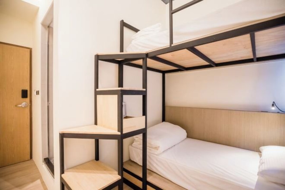 A bunk bed with a wooden frame and white bedding, a wooden door, and a light switch on the wall.