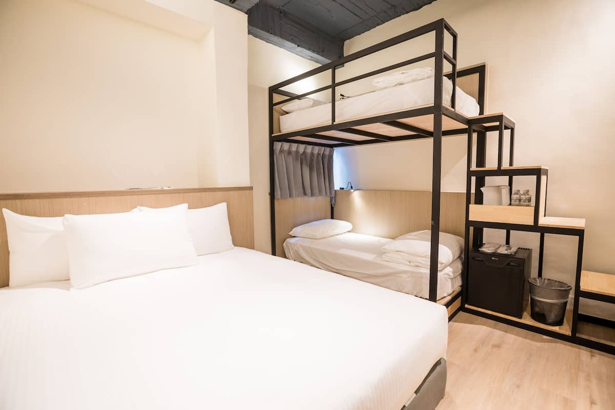 Una cama litera con estructura de madera, una mesita de noche con lámpara y un cubo de basura.