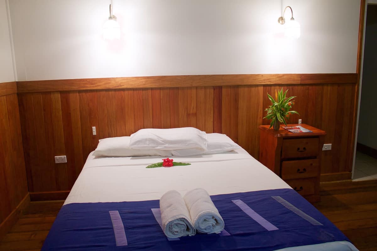 Un lit bien fait, avec des draps blancs, une couvre-lit bleue et deux serviettes enroulées. On y trouve une table de chevet en bois, sur laquelle sont posés une plante et un magazine.