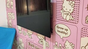 A room with a pink wall featuring Hello Kitty and other cartoon characters, a flat-screen TV mounted on the wall, a blue chair, and a small white table with a remote control. - Sleep Taipei Hotel Nan-Ya Branch (New Taipei City)