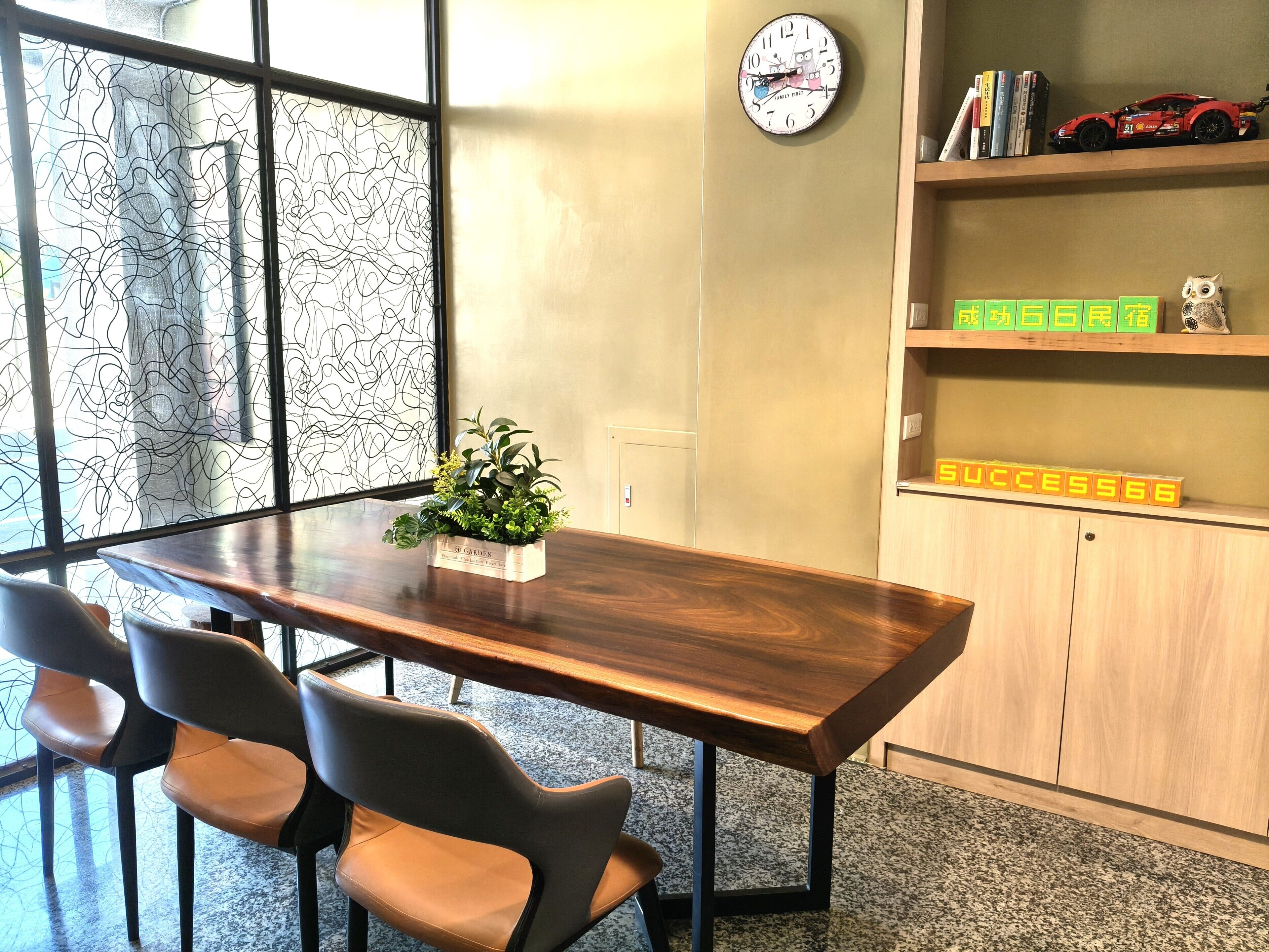 A conference room with a wooden table, four chairs, a clock, a bookshelf with books and a toy car, and a decorative plant.