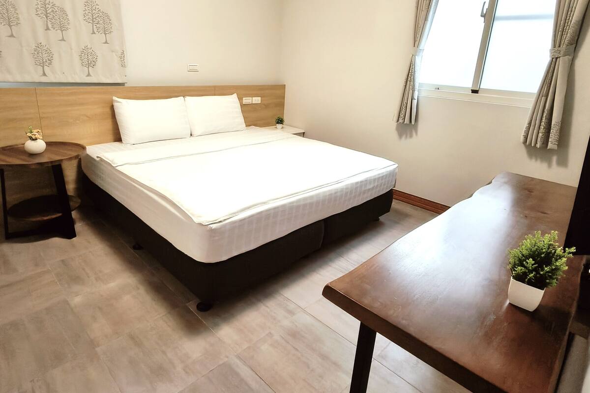 Un dormitorio con cama, mesitas de noche, una planta pequeña y una ventana con cortinas.