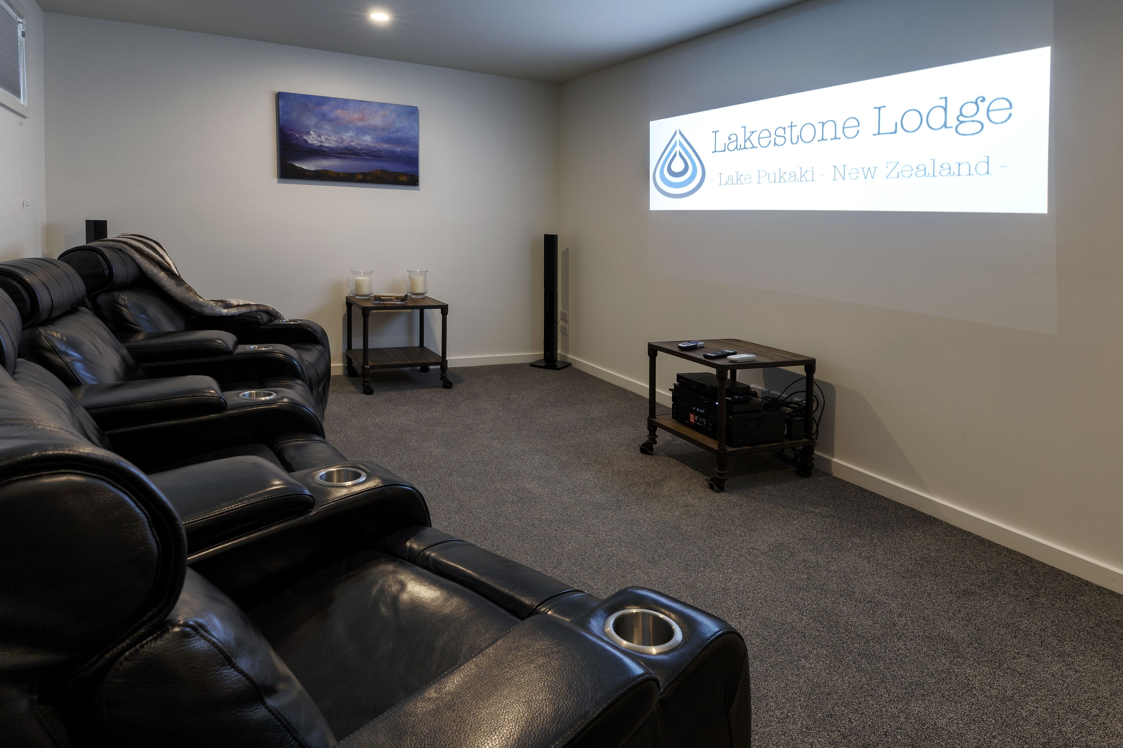 A massage therapy room with black leather chairs, a small table with a candle, and a projector screen displaying the name and location of Lakestone Lodge in New Zealand.