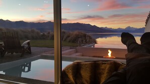 Una habitación con un ventanal amplio que da a un lago y montañas, un hogar y vistas a una terraza con mobiliario exterior.
