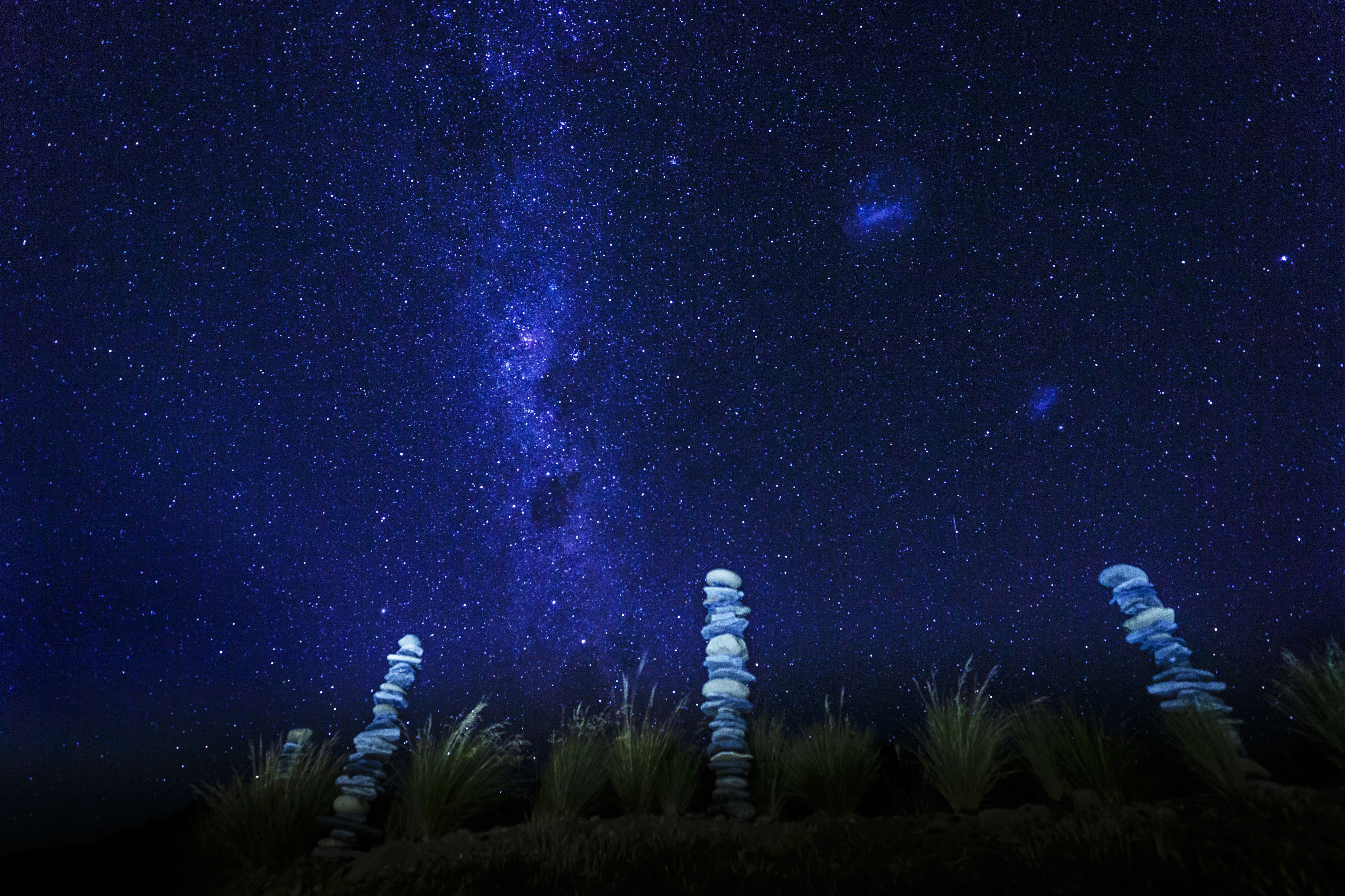 A night sky with a visible Milky Way and three stone stacks in a natural setting.