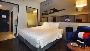 A hotel room with a bed, a seating area with a table, a TV, and a wall with a car and text. - Cabin Hotel (Jakarta)