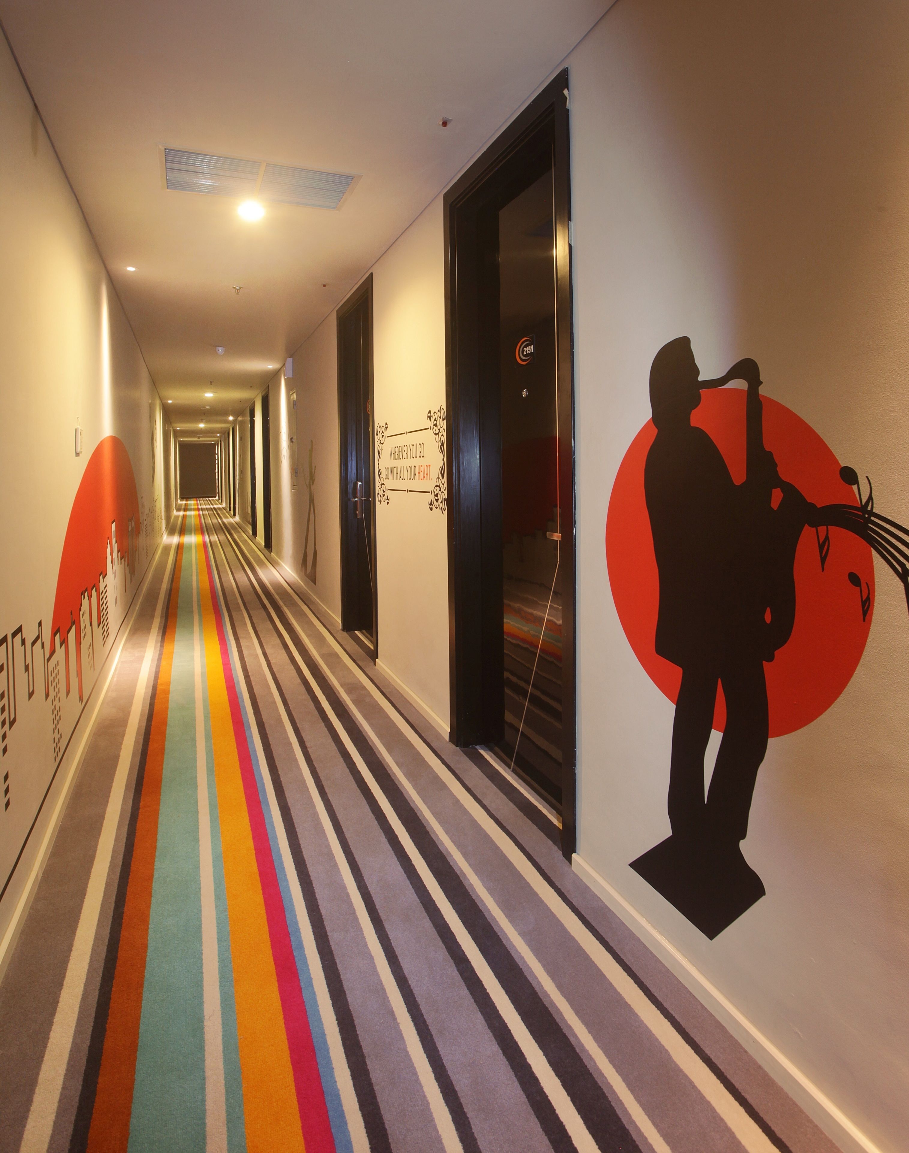 A hotel hallway with a colorful striped carpet, a wall mural of a saxophonist, and doors with a sign indicating room numbers.