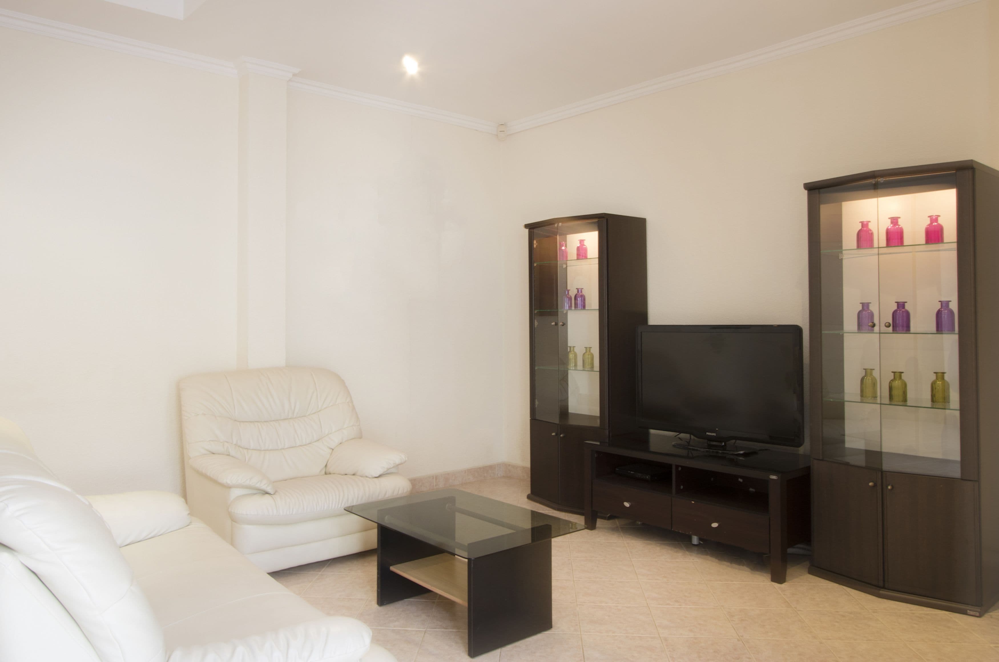 Uma sala de estar com sofá branco, mesa de centro de vidro, painel de entretenimento escuro com televisão de tela plana e dois armários de vidro com garrafas.