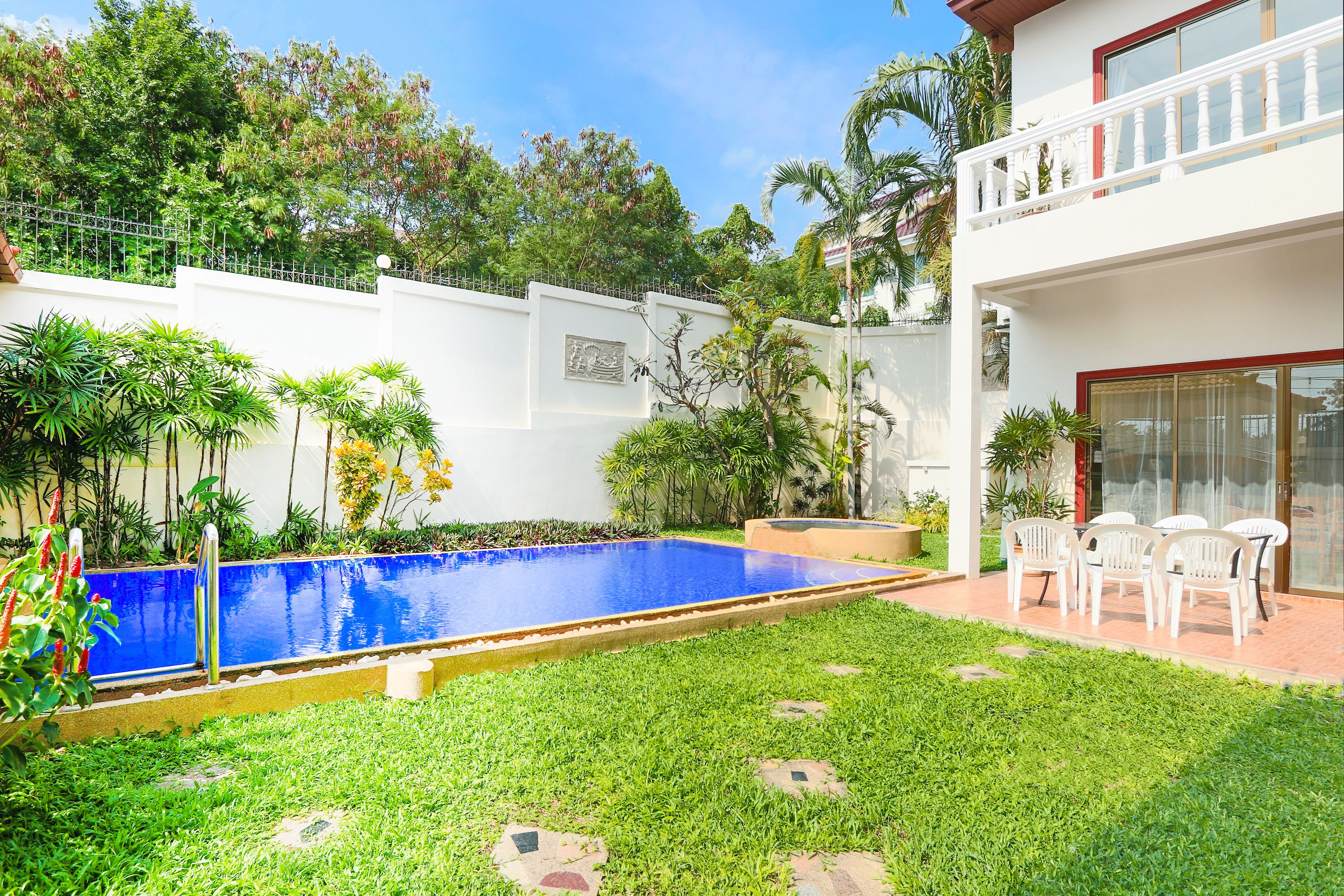A swimming pool surrounded by a well-maintained lawn, a white wall with a decorative panel, and a balcony with outdoor seating.