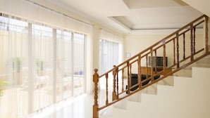 A spacious living room with a wooden staircase, large windows with sheer curtains, and a flat-screen TV on a stand. - Avoca Pool Villas (Pattaya)