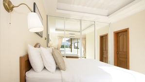 A bedroom with a bed, a wall-mounted lamp, a mirror, and a wooden door. - Avoca Pool Villas (Pattaya)