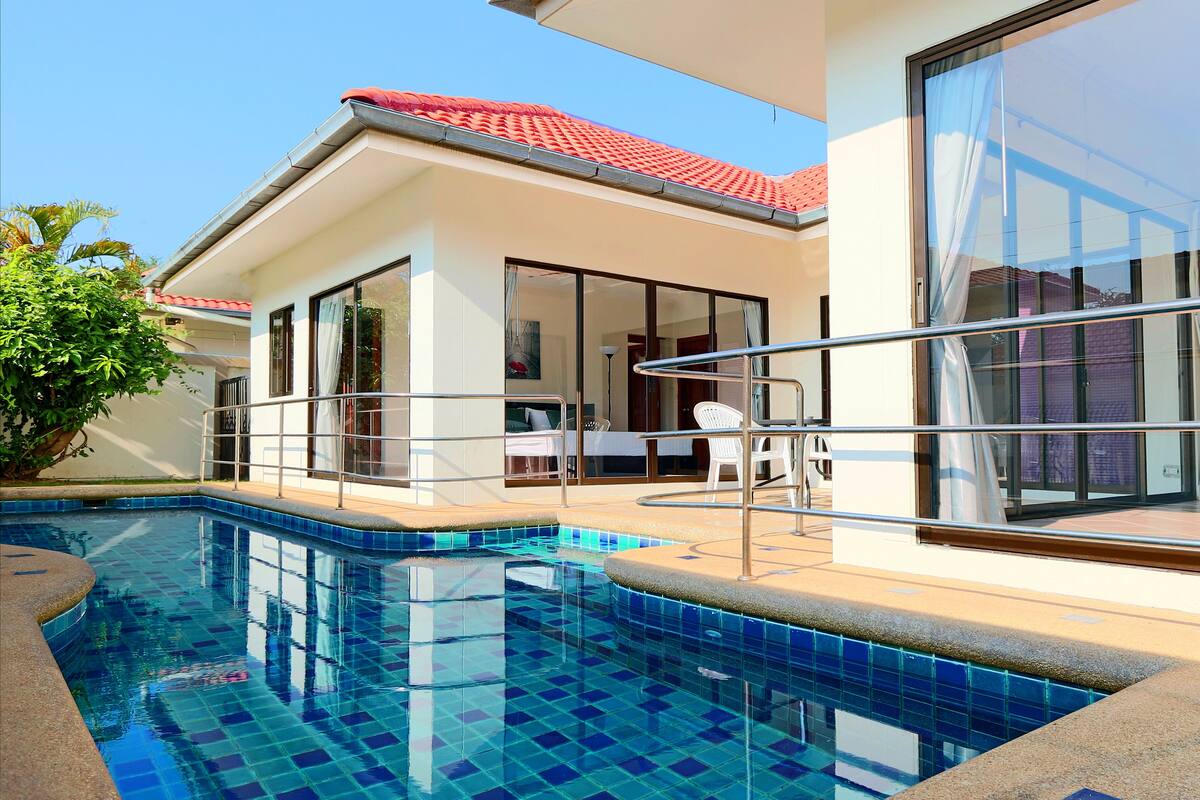 A swimming pool with clear blue tiles, a red-tiled roof, and a glass-enclosed balcony with a white chair.