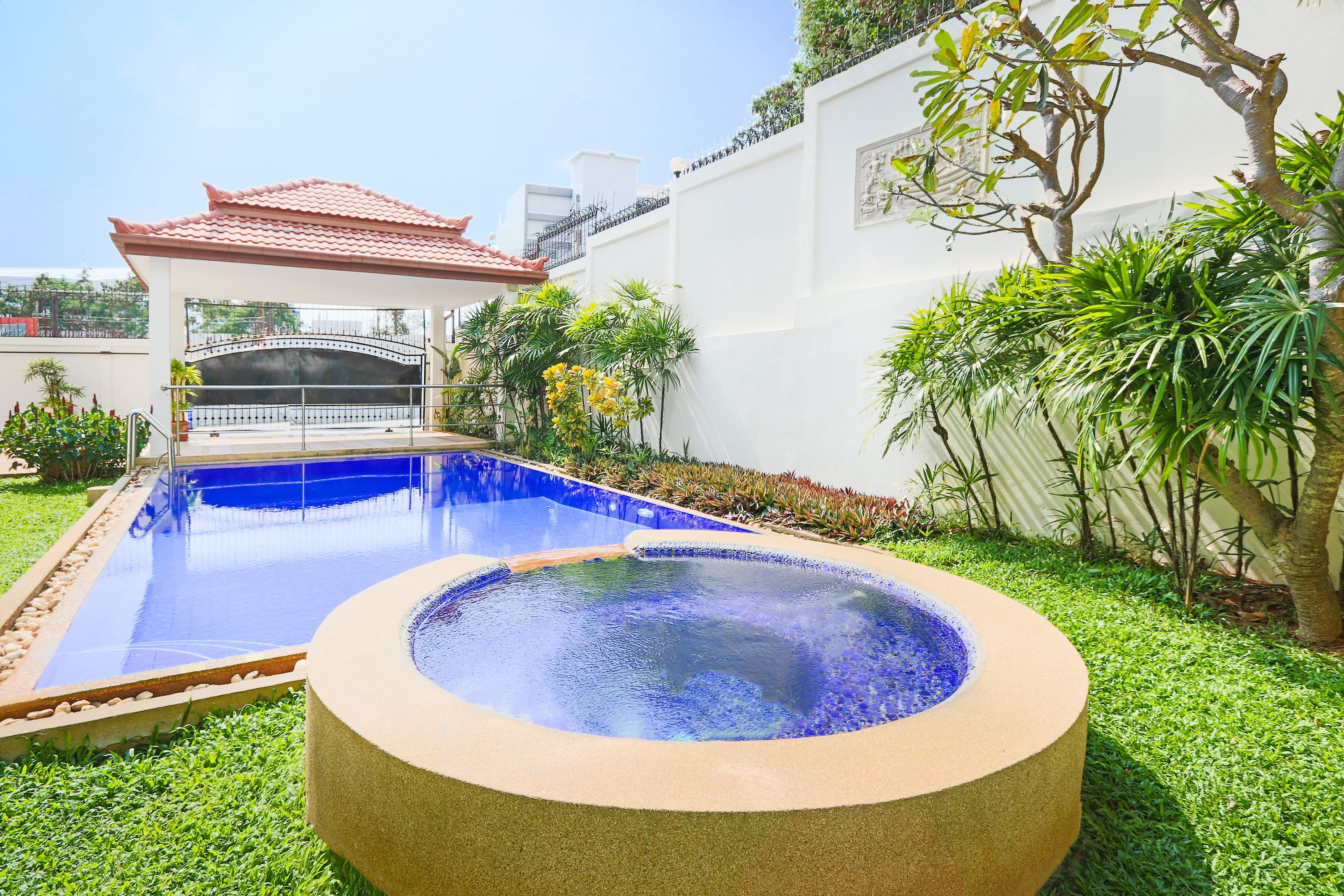 A swimming pool with a tiled roof structure, surrounded by well-maintained grass and various plants.