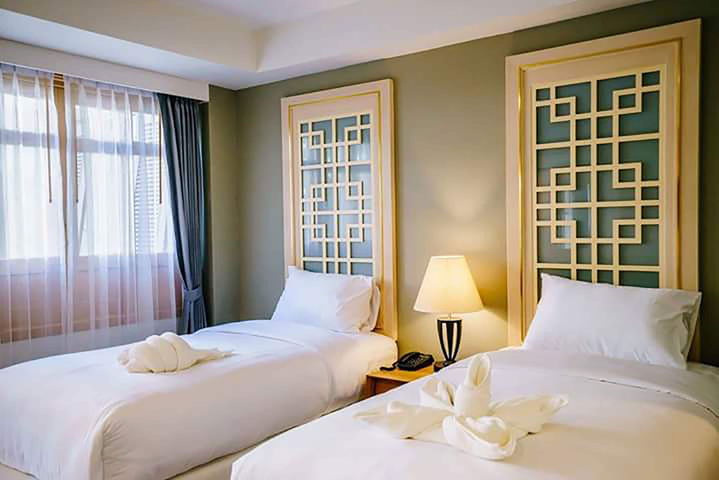 A hotel room with two beds, a window with sheer curtains, a wall-mounted lamp, and a decorative screen.