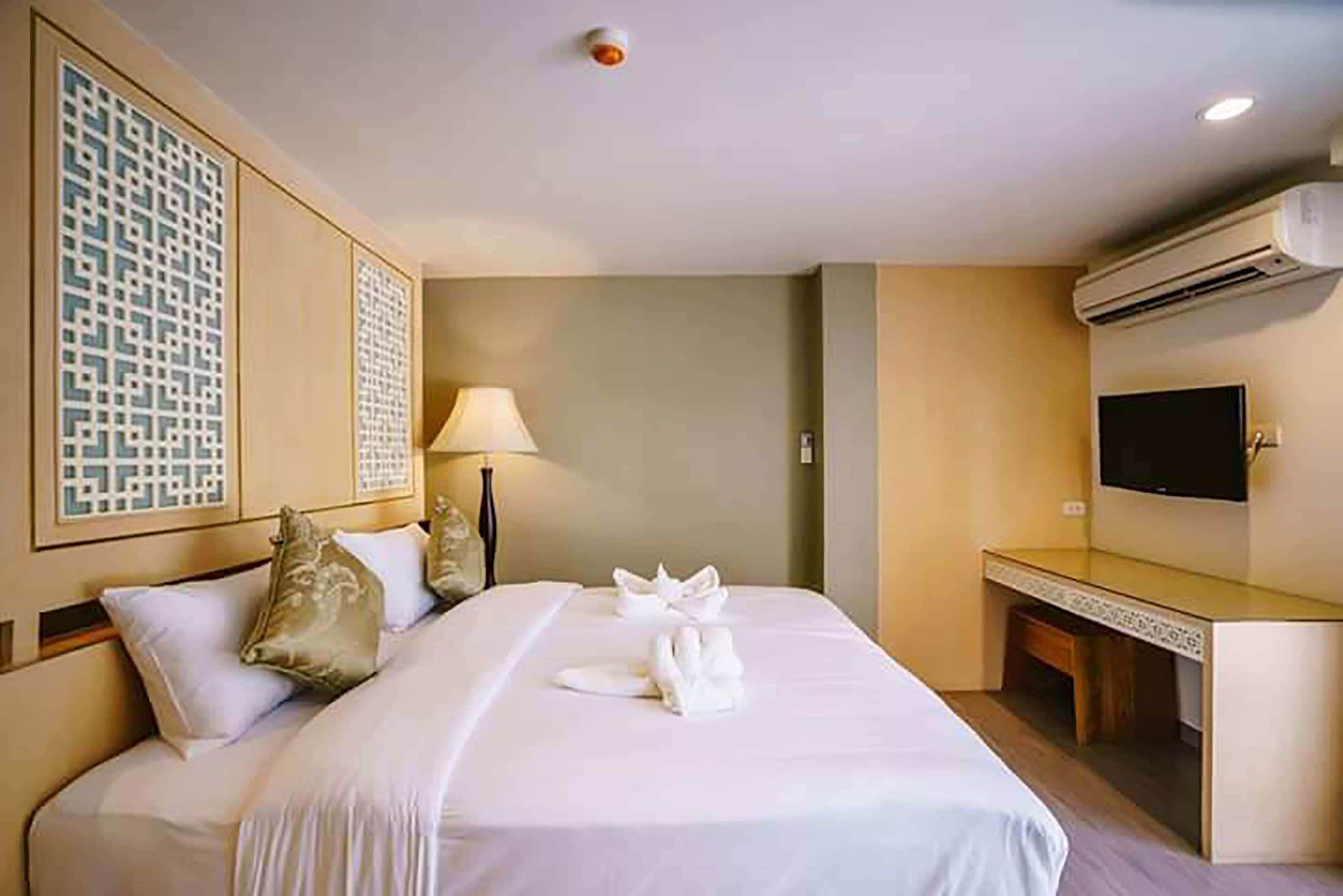 A hotel room with a neatly made bed, a bedside lamp, a flat-screen TV mounted on the wall, and a small desk with a chair.