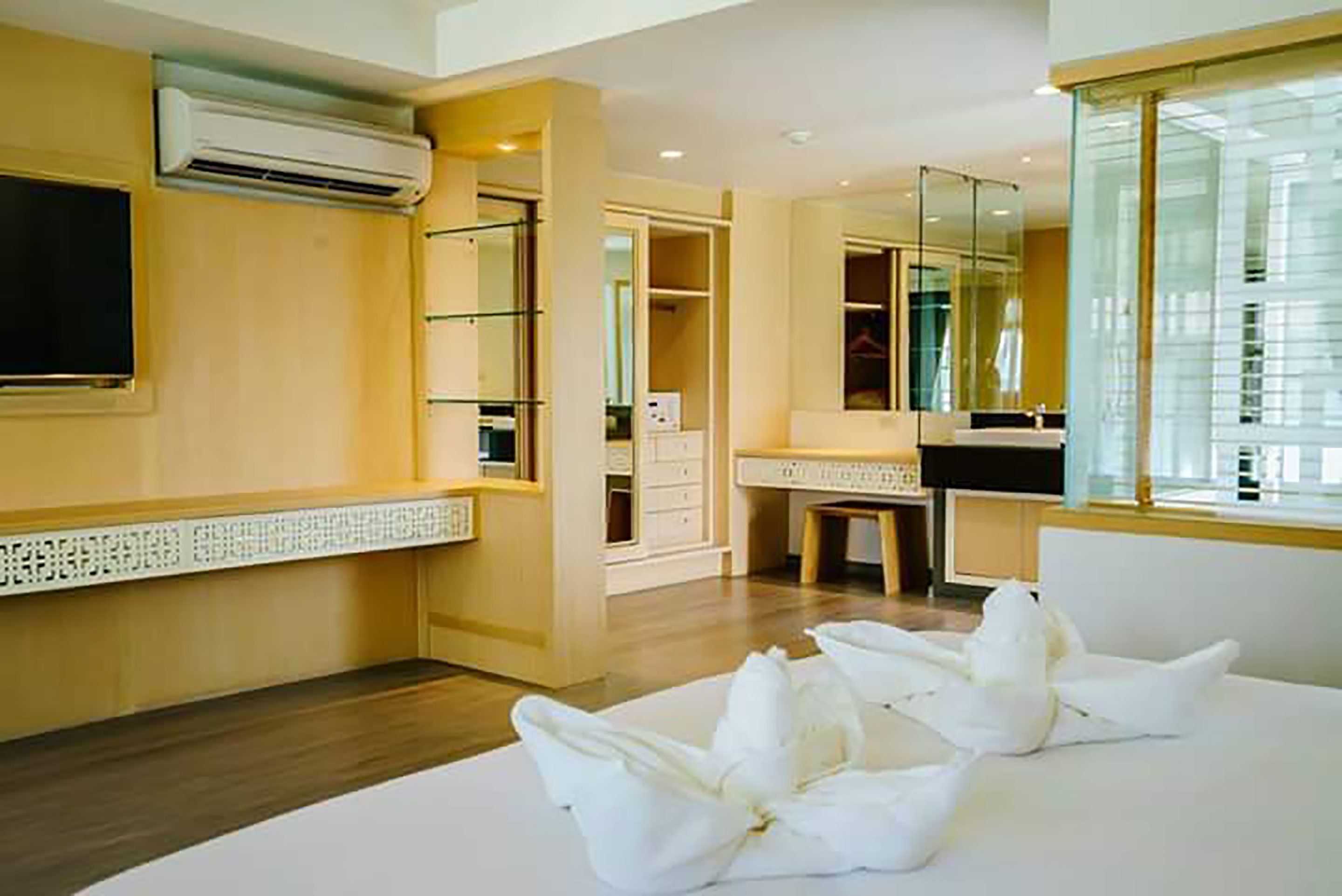 A hotel room with a wooden floor, a flat-screen TV mounted on the wall, a large mirror, and a white bed with decorative pillows.