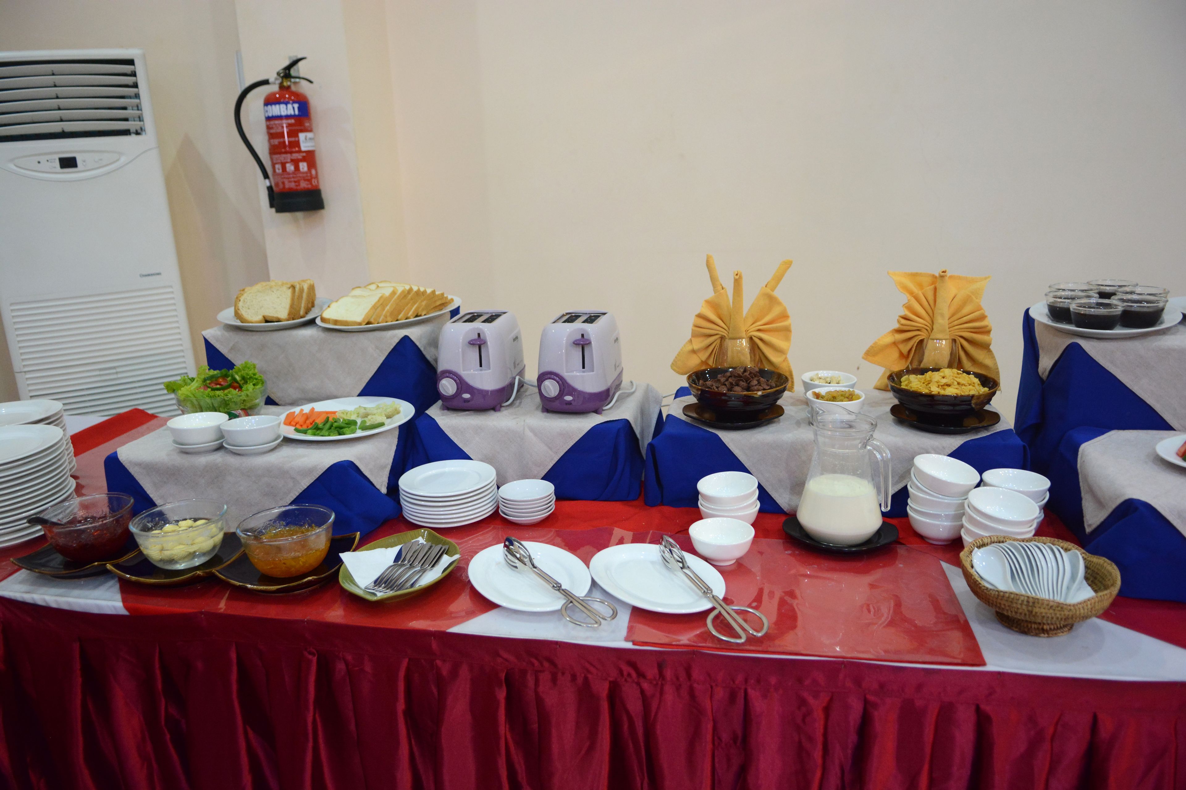 A buffet table with various food items and utensils, a fire extinguisher on the wall, and a stack of plates.