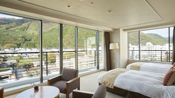 A hotel room with a large window offering a view of a city and mountains. There is a bed, a chair, and a small table.