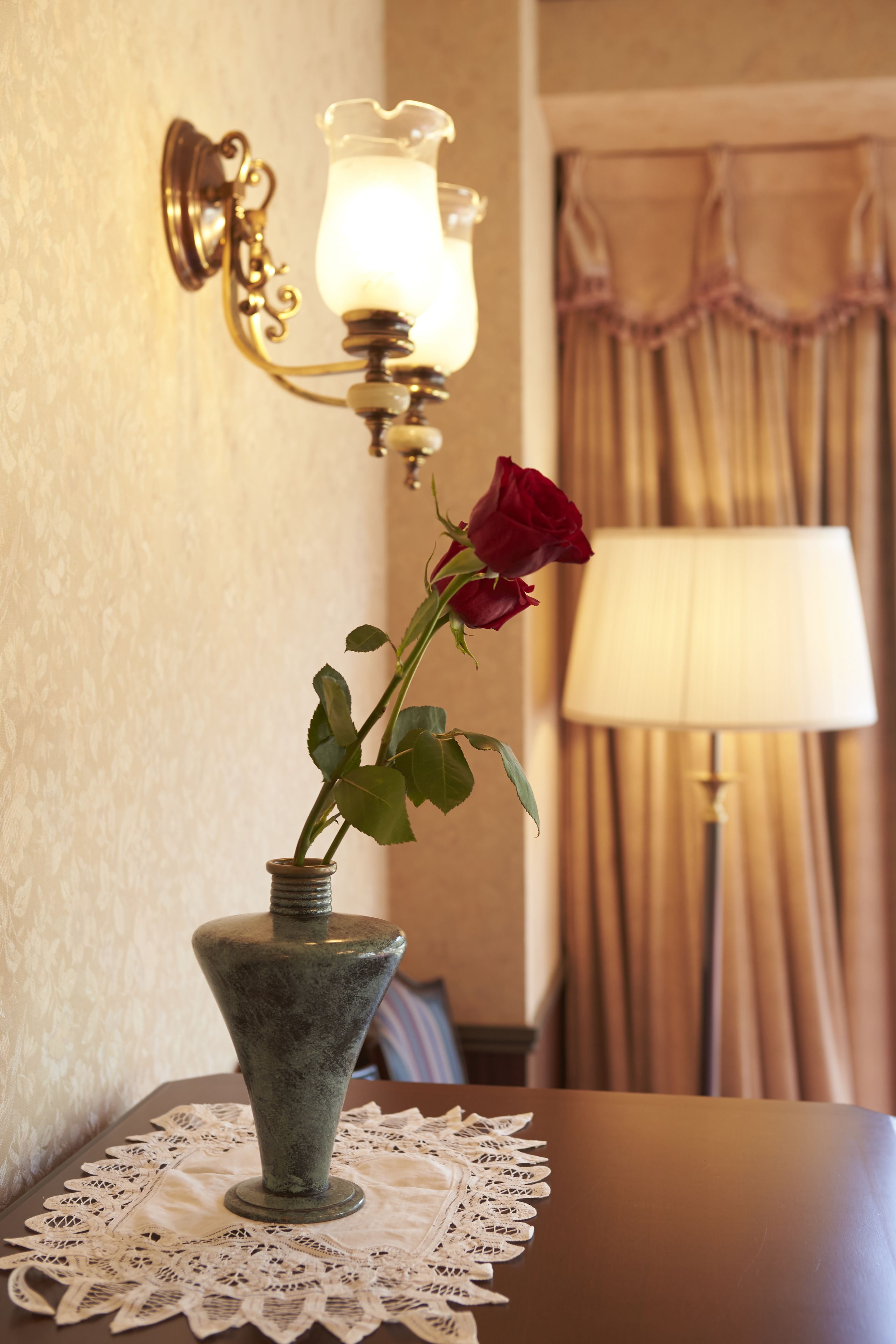 A vase with a single rose on a table, a wall-mounted lamp, and a curtain in the background.