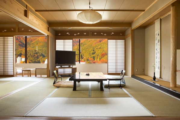 Una habitación japonesa tradicional con suelo de tatami, una mesa baja de tatami, un televisor y vistas del follaje otoñal a través de grandes ventanales.