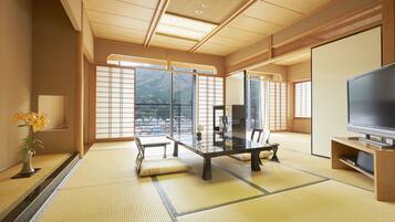 Una habitación japonesa tradicional con suelo de tatami, una mesa baja, un sofá, un televisor y vistas a montañas a través de amplios ventanales.