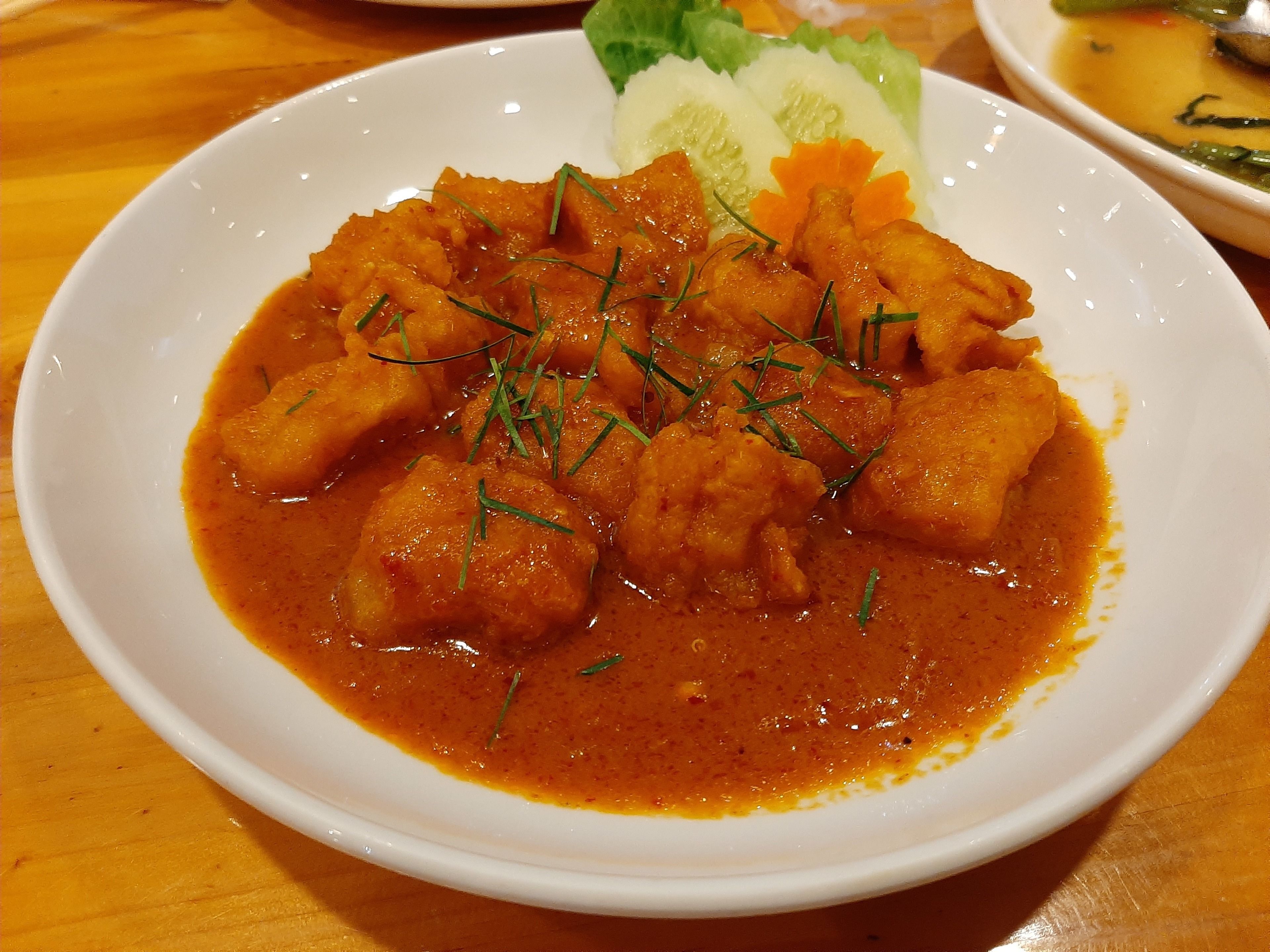 Un plato blanco con un guiso de curry anaranjado adornado con hierbas verdes, sobre una mesa de madera.